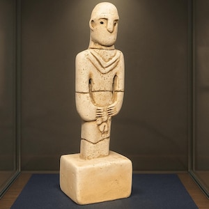 May include: A beige, stylized human figure sculpture. The figure has a simple, geometric design with a rectangular base. The arms are bent, and the hands are holding an object. The sculpture is displayed in a glass case.