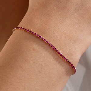 May include: A slender gold bracelet set with many small, bright red gemstones. The bracelet is fine and rests gently on the wrist, catching the light. The jewellery is shown against a soft, neutral background.