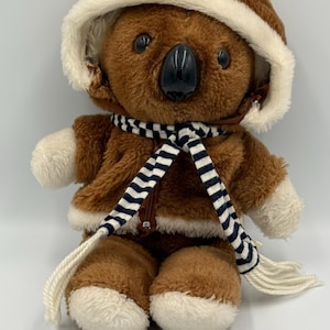 Puede incluir: Un oso koala de peluche con una chaqueta con capucha marrón con ribete color crema y una bufanda a rayas blancas y negras. El koala tiene una nariz negra y patas y pies color crema. El peluche está sentado.
