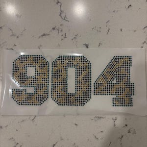 May include: A rectangular iron-on transfer sheet featuring the number 904 created with small rhinestones. The digits are filled with a mix of gold and dark teal rhinestones, arranged in a block-style font on a clear adhesive backing material.