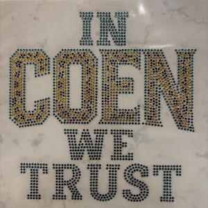 May include: A rhinestone transfer sheet featuring the text IN COEN WE TRUST. The word COEN is filled with a leopard print pattern made of gold and black rhinestones, while the words IN, WE, and TRUST are composed of solid dark blue rhinestones.