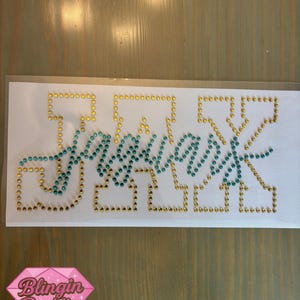 May include: A rhinestone transfer with the word "Yessss" in teal and gold rhinestones. The word is outlined in gold rhinestones. The image also includes a pink diamond logo with the words "Blingin' Baddies".