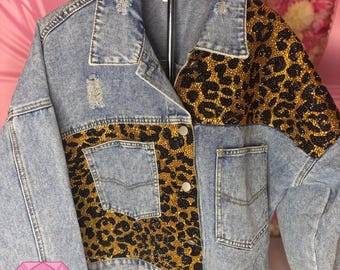 Op de afbeelding: Lichtblauwe spijkerjas met luipaardprint details. De jas heeft een klassieke kraag, knoopsluiting en twee voorzakken. De luipaardprint is versierd met zwarte en gouden strass-steentjes. De jas heeft distressed details en de tekst "Blingin' Baddies".