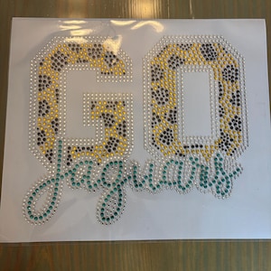 Puede incluir: Una hoja blanca con las palabras "GO JAGUARS" en pedrería. Las letras "GO" están rellenas con un estampado de leopardo amarillo y negro. La palabra "Jaguars" está escrita en una fuente cursiva turquesa.