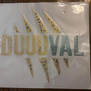 May include: A rhinestone transfer sheet featuring the word DUUVAL in gold and teal crystals. The design includes gold and dark blue claw mark accents above and below the text, all arranged on a clear, flexible adhesive backing for fabric application.