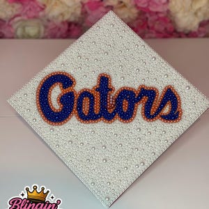 Könnte beinhalten: Eine weiße Abschlusskappe, verziert mit kleinen weißen Perlen. Das Wort "Gators" ist in Blau und Orange geschrieben und mit kleinen Perlen umrandet. Das Blingin' Baddies-Logo befindet sich in der unteren linken Ecke.
