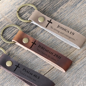 Puede incluir: Tres llaveros de cuero en color beige, marrón y marrón oscuro, cada uno con un anillo de metal y un diseño de cruz. Los llaveros tienen texto grabado de la Biblia: Josué 1:9, Jeremías 29:11 y Salmo 46:5.
