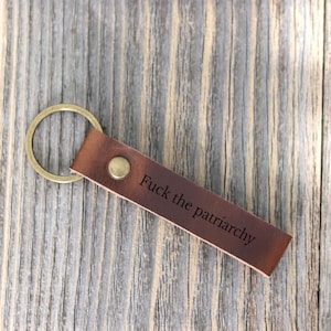 May include: A brown leather keychain with a brass ring and a brass rivet. The text "Fuck the patriarchy" is engraved on the leather. The keychain is lying on a weathered wooden surface.