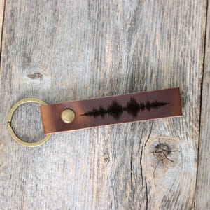 May include: A brown leather keychain with a gold-colored ring. The leather strap features a sound wave design and a brass-colored button. The keychain is displayed on a weathered wooden surface.