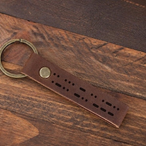 Op de afbeelding: Een donkerbruine leren sleutelhanger met een reeks lasergesneden morsecode-stippen en streepjes. De leren riem is vastgezet met een ronde, messingkleurige metalen klinknagel en bevestigd aan een bijpassende ronde metalen sleutelring.