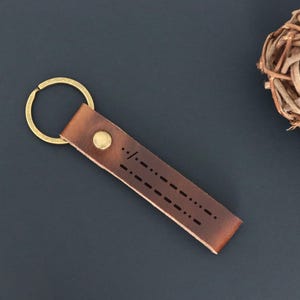 Peut inclure: Un porte-clés en cuir marron avec un anneau et un rivet de couleur or. La lanière en cuir présente une série de perforations en pointillés et en tirets. Le porte-clés est posé sur une surface bleu foncé, avec des sphères décoratives tressées en arrière-plan.