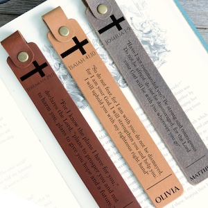 Puede incluir: Tres marcapáginas rectangulares de cuero en marrón oscuro, tostado y gris. Cada uno tiene un botón metálico, un gráfico de cruz negra y versículos bíblicos impresos: Jeremías 29:11, Isaías 41:10 y Josué 1:9. El nombre Olivia aparece en uno.