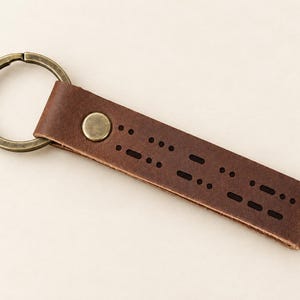 May include: A dark brown leather keychain featuring a rectangular strap with laser-cut Morse code patterns. The leather is secured by a circular antique brass rivet and attached to a matching round antique brass split key ring.