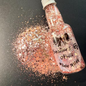 May include: A clear bottle filled with pink glitter, with a silver cap, and a black bow graphic. The bottle is labeled "Creations By Holli Pixie Alloy". A pile of glitter spills out onto a black surface.