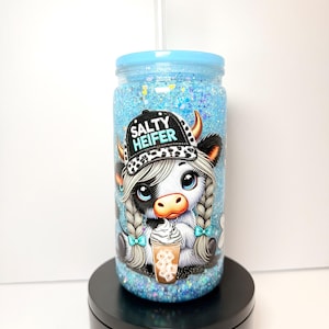 May include: A blue glitter tumbler with a cow graphic wearing a hat that says "Salty Heifer". The tumbler has a light blue lid and a clear straw. The cow graphic has braids and is holding a coffee drink.