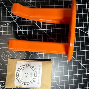 The orange garage hook and hardware kit placed on a grey measurement grid to show the physical scale and size comparison.