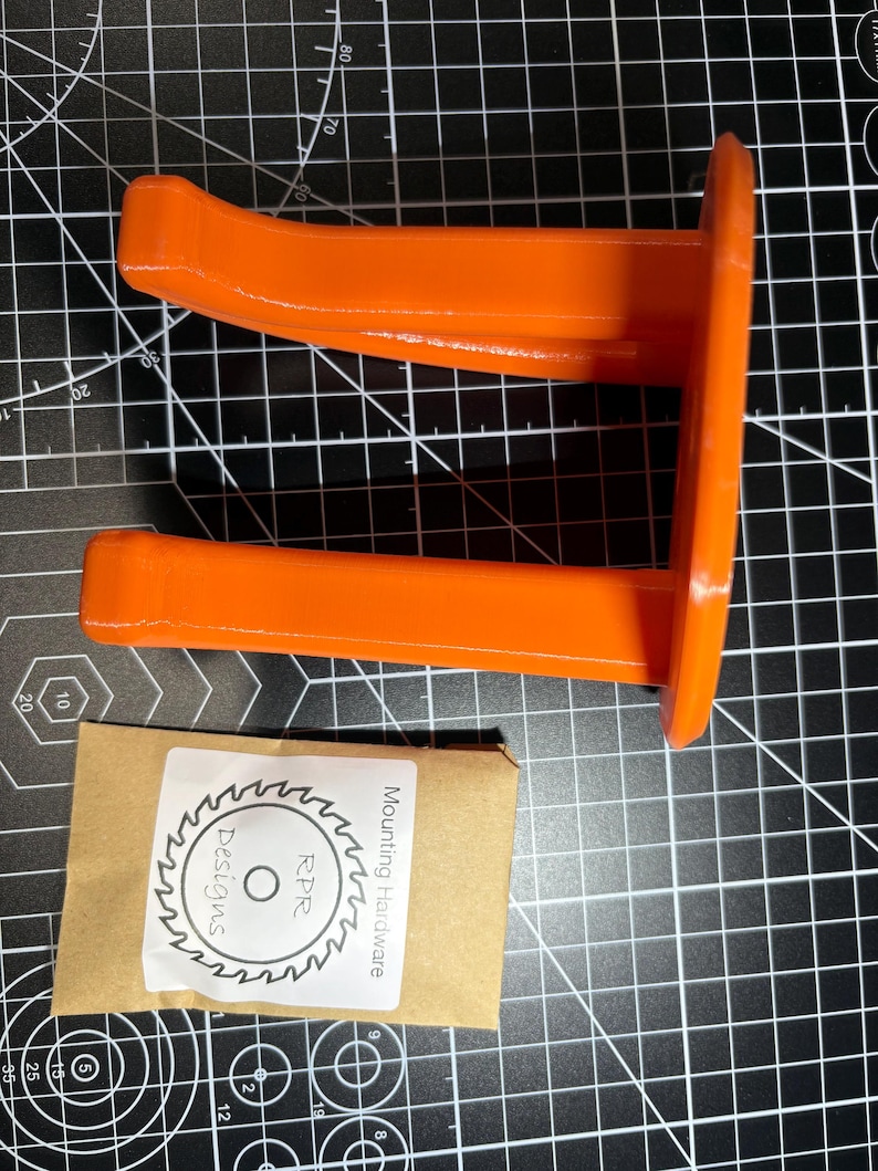 The orange garage hook and hardware kit placed on a grey measurement grid to show the physical scale and size comparison.