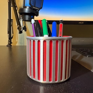A silver ribbed pencil holder filled with colorful pens and markers on a dark wooden desk in a gaming setup, positioned in front of a computer monitor and professional microphone.