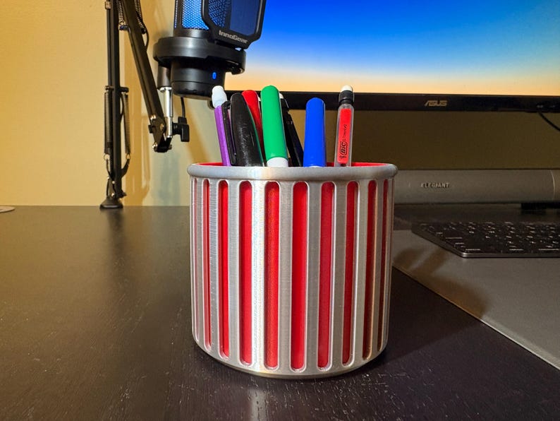 A silver ribbed pencil holder filled with colorful pens and markers on a dark wooden desk in a gaming setup, positioned in front of a computer monitor and professional microphone.