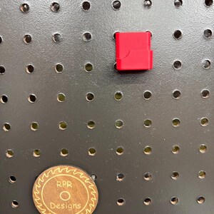 An empty red RPR Designs caliper holder mounted on a pegboard next to a circular wooden brand coin for scale.