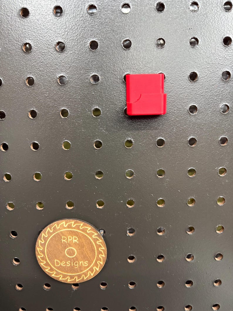 An empty red RPR Designs caliper holder mounted on a pegboard next to a circular wooden brand coin for scale.