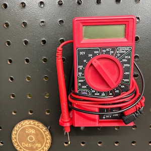 May include: A red digital multimeter with black and red wires and probes. The multimeter has a digital display and a rotary dial with various settings. A wooden disc with the text "RPR Designs" is also visible.