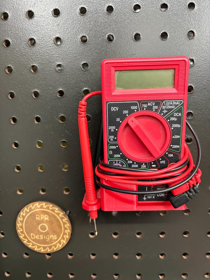 A front view of a red pegboard-mounted multimeter holder securely cradling a digital meter with test leads neatly wrapped and stored in integrated side loops.