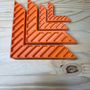 May include: An assortment of bright orange corner clamps, arranged on a wooden surface. The clamps are L-shaped, with a series of diagonal grooves. The sizes vary, suggesting a set for different project needs. The tools are designed for woodworking or construction.