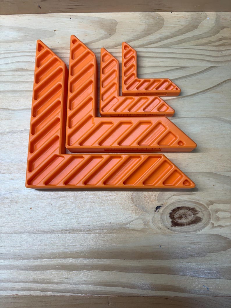 May include: An assortment of bright orange corner clamps, arranged on a wooden surface. The clamps are L-shaped, with a series of diagonal grooves. The sizes vary, suggesting a set for different project needs. The tools are designed for woodworking or construction.