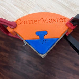 May include: An orange and blue CornerMaster corner clamping tool with the text CornerMaster and 1/2 inch printed on the surface. The tool is secured to a wooden board with red and black clamps, designed to hold a 90-degree corner in place.