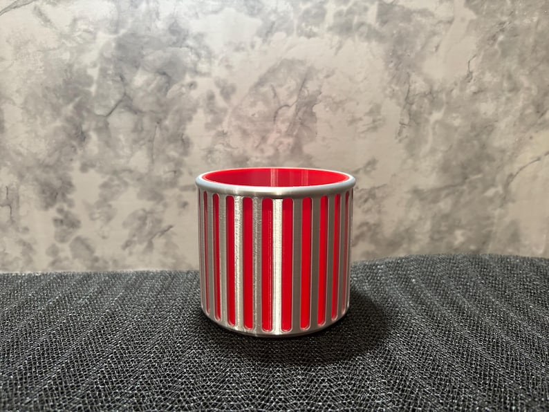 A futuristic silver pencil holder with a vibrant red interior and vertical slotted ribs on a grey textured surface.