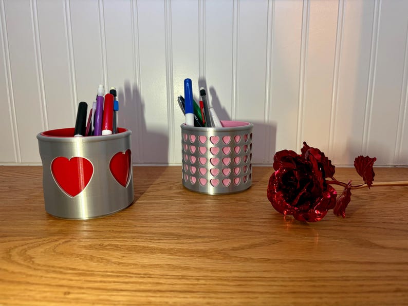 Two silver 3D-printed heart pencil holders with red and pink interiors sitting on a wooden desk next to a metallic red rose and pens.