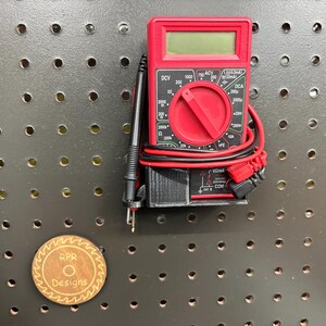 A professional black multimeter holder mounted on a pegboard, shown with a red digital tester and test leads neatly coiled using the integrated side loops.