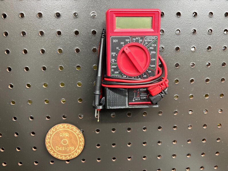 A professional black multimeter holder mounted on a pegboard, shown with a red digital tester and test leads neatly coiled using the integrated side loops.