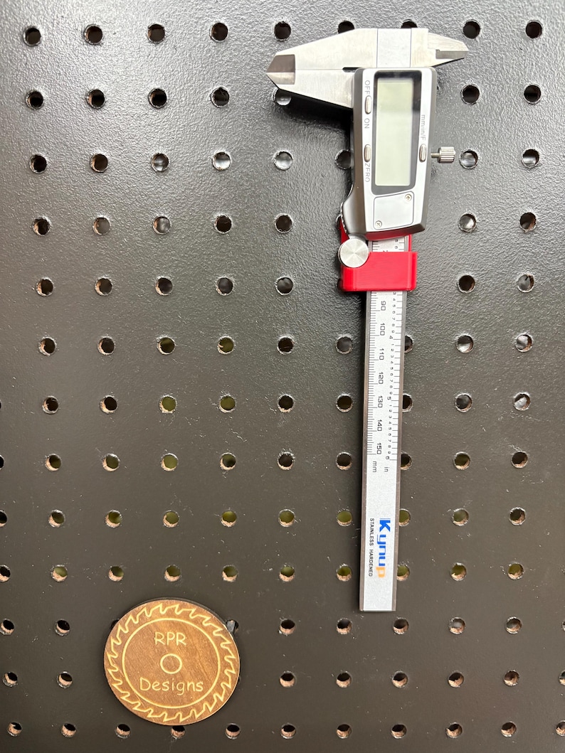 A 6-inch digital caliper securely resting in a bright red 3D-printed RPR Designs holder on a black metal pegboard.