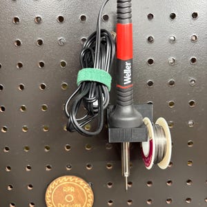 Black 3D printed soldering iron cradle mounted on a dark pegboard holding a Weller iron and a spool of solder for one-handed use.