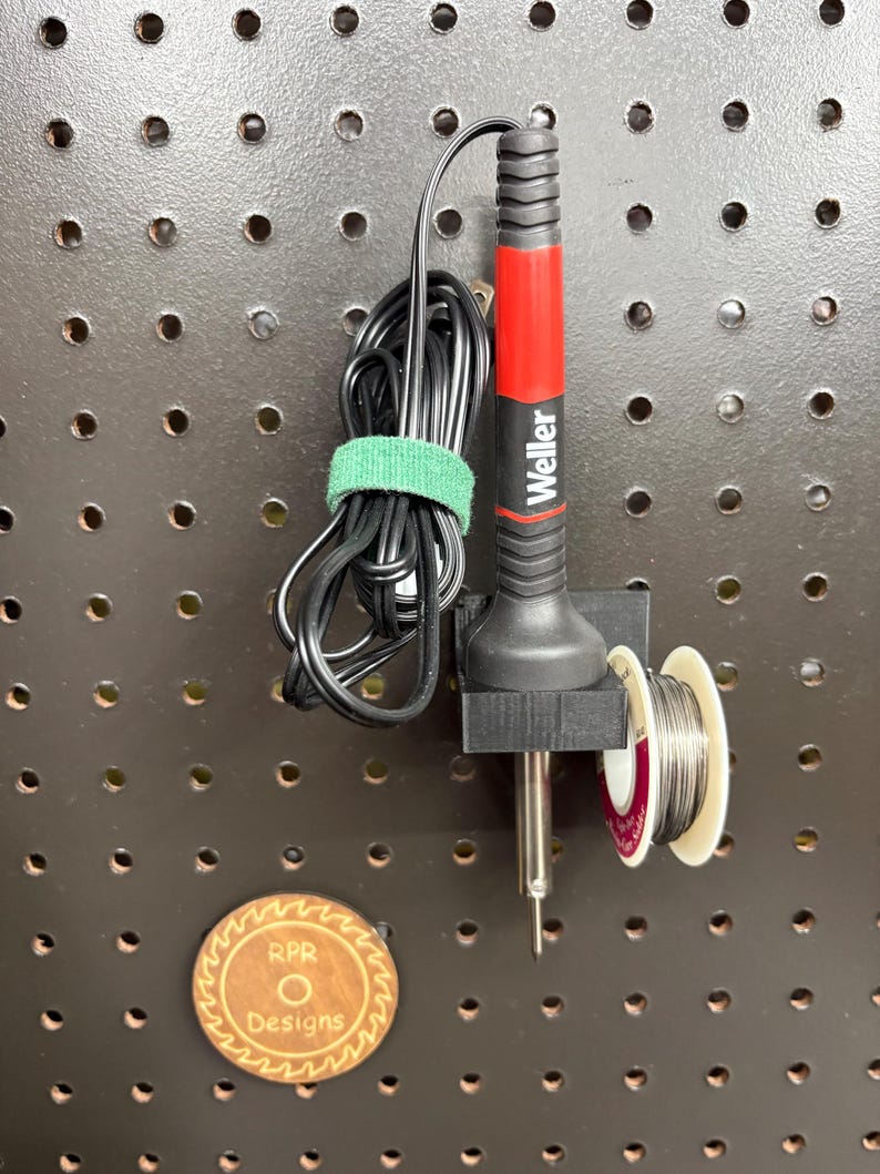Black 3D printed soldering iron cradle mounted on a dark pegboard holding a Weller iron and a spool of solder for one-handed use.