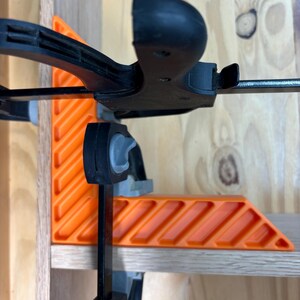 May include: An orange and black corner clamp is used to hold two pieces of wood together. The clamp has an L-shaped orange body with a series of angled slots and a black handle. The clamp is attached to a wooden shelf.