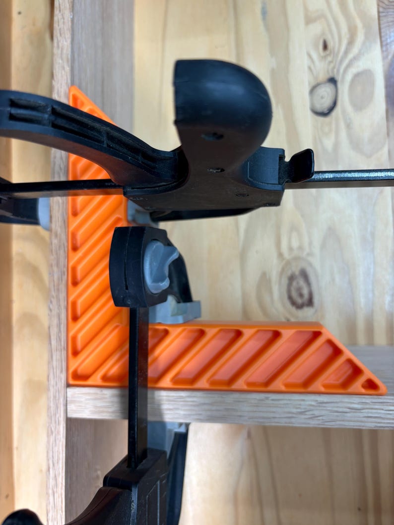 May include: An orange and black corner clamp is used to hold two pieces of wood together. The clamp has an L-shaped orange body with a series of angled slots and a black handle. The clamp is attached to a wooden shelf.