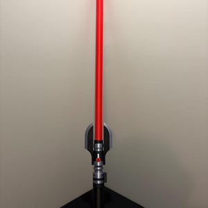 May include: A red lightsaber prop with a black and silver hilt. The lightsaber has a long, cylindrical red blade and a detailed handle with silver and black accents. The prop is designed to resemble a lightsaber from a science fiction franchise.