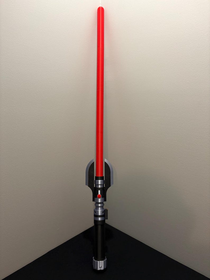May include: A red lightsaber prop with a black and silver hilt. The lightsaber has a long, cylindrical red blade and a detailed handle with silver and black accents. The prop is designed to resemble a lightsaber from a science fiction franchise.