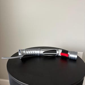 Puede incluir: Un accesorio de sable de luz plateado y negro con un bot&oacute;n rojo y una hoja curva. El accesorio est&aacute; sobre una superficie redonda y negra. El sable de luz tiene un dise&ntilde;o met&aacute;lico detallado.