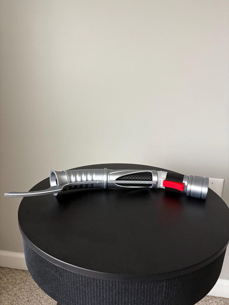 Puede incluir: Un accesorio de sable de luz plateado y negro con un bot&oacute;n rojo y una hoja curva. El accesorio est&aacute; sobre una superficie redonda y negra. El sable de luz tiene un dise&ntilde;o met&aacute;lico detallado.