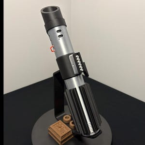 May include: A detailed replica of a lightsaber, featuring a black and silver handle with intricate detailing. The lightsaber is displayed on a black stand with a small, brown box. The overall design is reminiscent of a popular science fiction franchise.