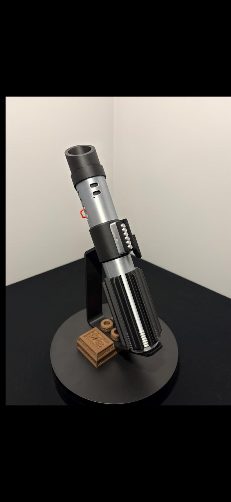 May include: A detailed replica of a lightsaber, featuring a black and silver handle with intricate detailing. The lightsaber is displayed on a black stand with a small, brown box. The overall design is reminiscent of a popular science fiction franchise.