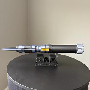 May include: A blue and black lightsaber prop with silver and yellow accents, resting on a black stand. The lightsaber has a long, thin blade and a textured handle. The stand is also black and has a detailed design.