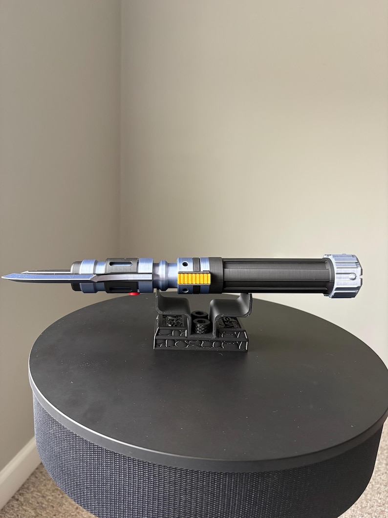 May include: A blue and black lightsaber prop with silver and yellow accents, resting on a black stand. The lightsaber has a long, thin blade and a textured handle. The stand is also black and has a detailed design.