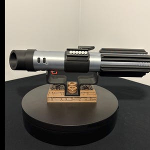 May include: A detailed replica of a lightsaber, featuring a black and silver handle with intricate detailing. The prop is displayed on a black stand with a small, decorative structure. The lightsaber's design is reminiscent of a popular science fiction franchise.