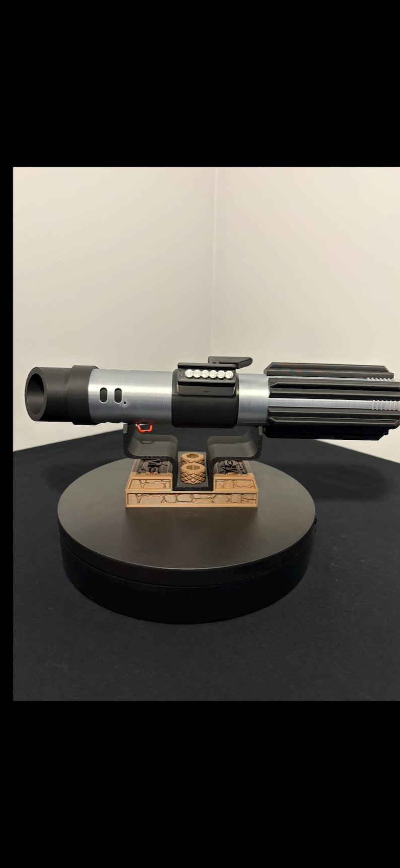 May include: A detailed replica of a lightsaber, featuring a black and silver handle with intricate detailing. The prop is displayed on a black stand with a small, decorative structure. The lightsaber's design is reminiscent of a popular science fiction franchise.
