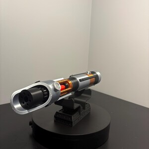 May include: A silver and black lightsaber prop with orange and red accents, displayed on a black stand. The prop features a detailed design with a cylindrical handle and a visible internal mechanism. The lightsaber is a replica of a popular science fiction weapon.